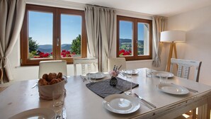 A dining table set with plates, cutlery, and a bowl of croissants, with a view of the countryside through large windows.