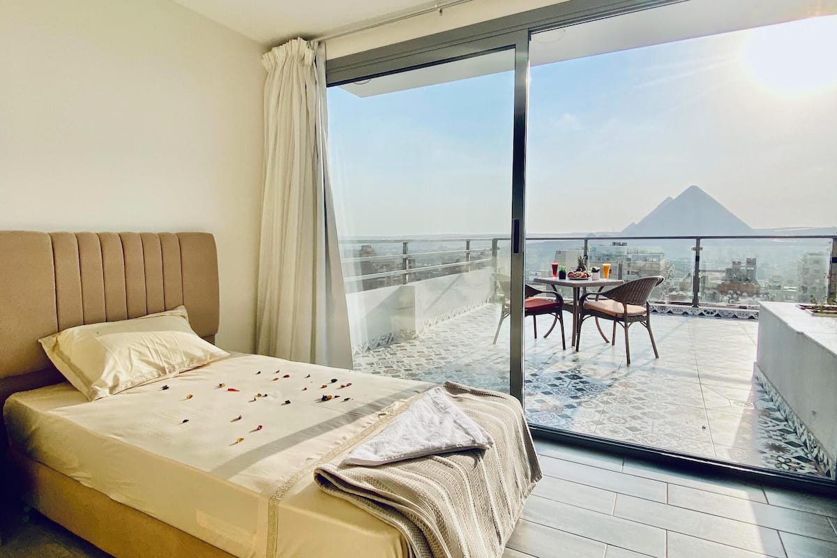A bedroom with a large bed, a view of a cityscape and a mountain through a sliding glass door, and a balcony with a table and chairs.