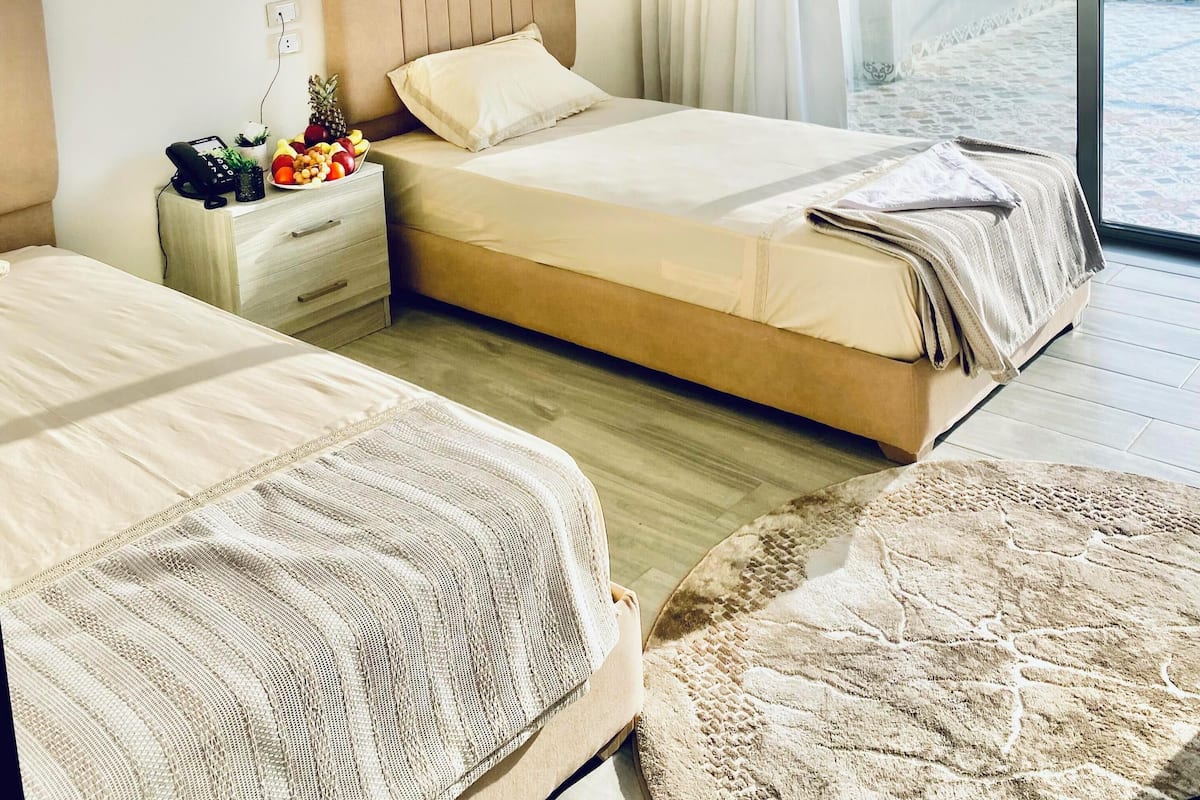 A hotel room with two beds, a bedside table with a fruit bowl, a phone, and a view of the city through large windows.