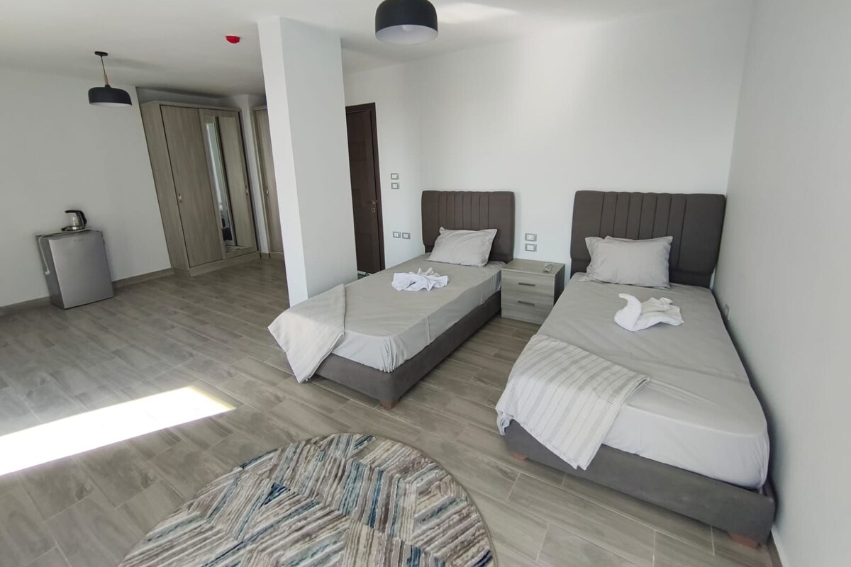 A modern hotel room with two beds, a small seating area, and a kitchenette.