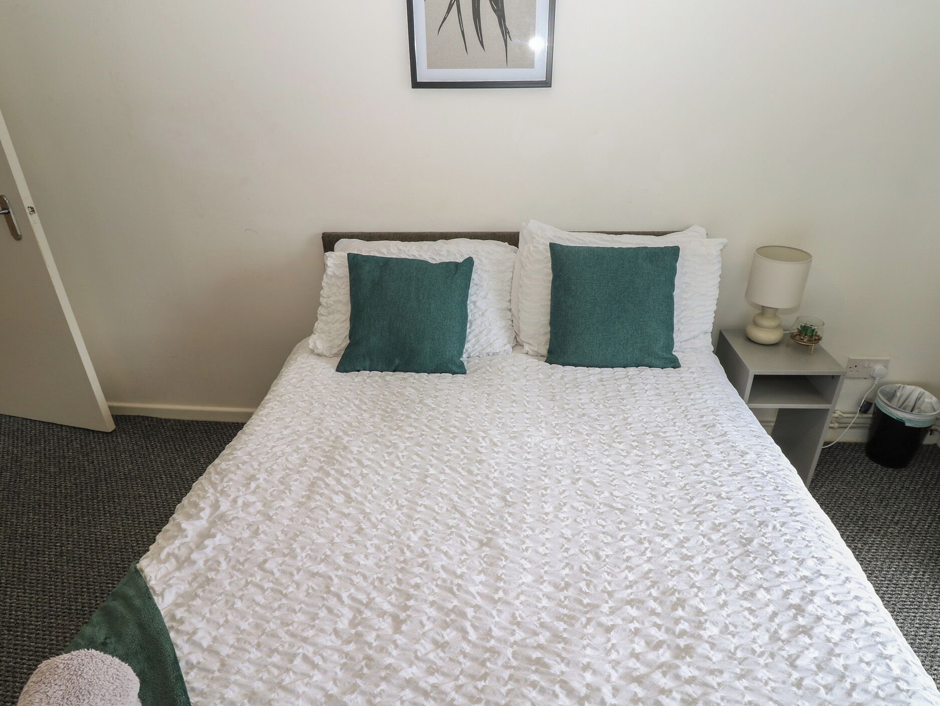 A neatly made bed with white bedding and two green pillows, a bedside table with a lamp and a small plant, and a framed picture on the wall.