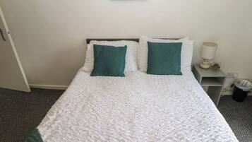 A neatly made bed with white bedding and two green pillows, a bedside table with a lamp and a small plant, and a framed picture on the wall.