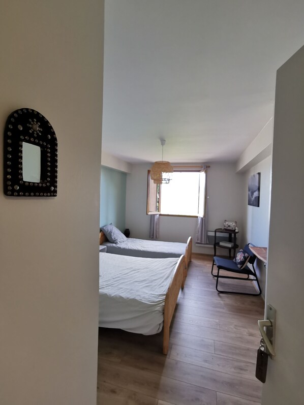 A room with two beds, a small table, a chair, a hanging light fixture, and a window. - SARL SCOP BIENVENUE A L AUBERGE (Sauze)