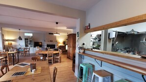 Un comedor amplio con mesas y sillas de madera, una zona de cocina con mostrador y taburetes de madera, y una chimenea al fondo.