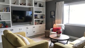 A living room with a large flat-screen TV, a sofa, a coffee table, and a bookshelf filled with decorative items. - Charming 2-bedroom house in lovely New Port Richey with pool. (New Port Richey)