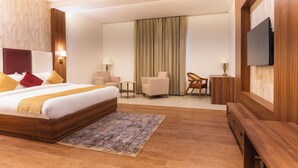 A hotel room with a large bed, a TV mounted on the wall, a small table, a chair, and a desk. - Haut Monde Rajputana Palace (Behror)