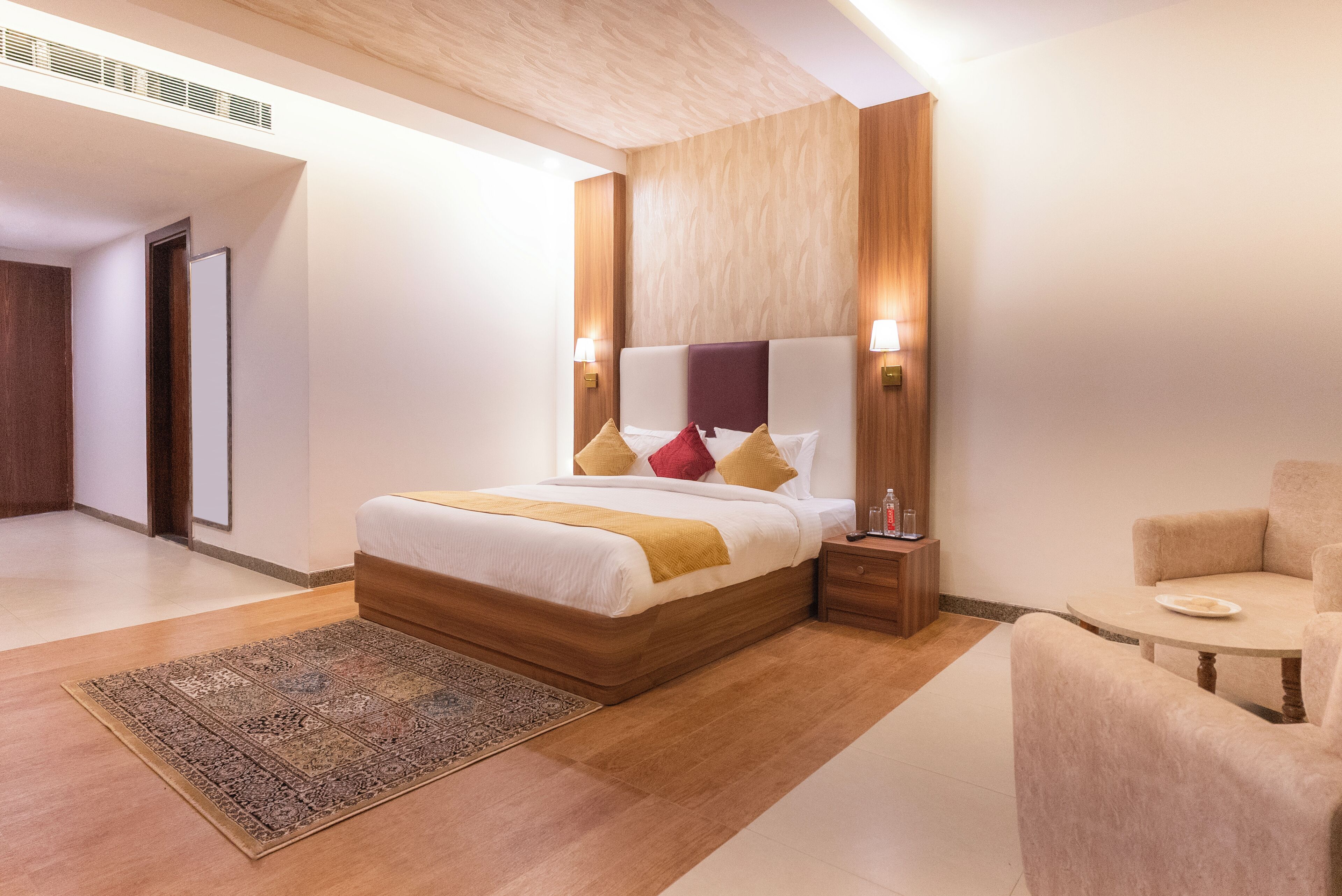 A modern hotel room with a large bed, a wooden headboard, bedside lamps, a small table, and a chair.