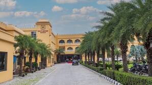 A hotel entrance with a paved driveway, palm trees, and a clear sky. - Haut Monde Rajputana Palace (Behror)
