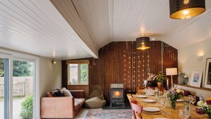 Ein gemütliches Wohnzimmer mit Kamin, eine Essecke mit Holztisch, gedeckt für eine Mahlzeit, und ein großes Fenster mit Blick ins Freie.