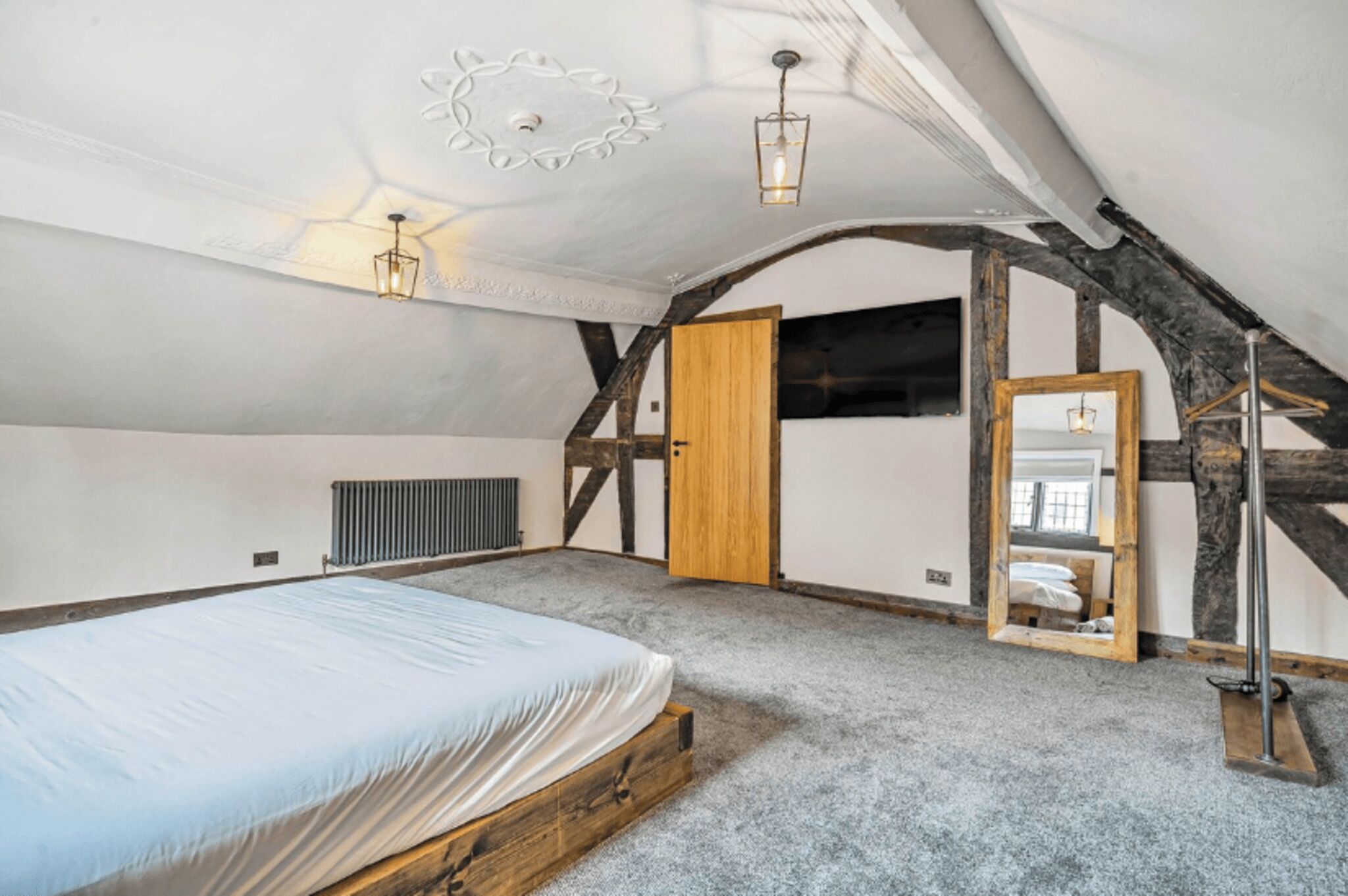 A bedroom with a large bed, a wooden door, a mirror, and a ceiling with exposed beams.