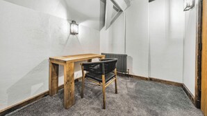 A small, narrow room with a wooden table and a black chair, a radiator, and a wall-mounted light fixture. - Chester Rose, Chester (Chester)