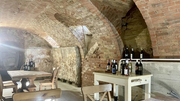 Una cantina vinicola sotterranea con volte a mattoni, tavoli e sedie in legno, e una varietà di bottiglie di vino esposte su un bancone.
