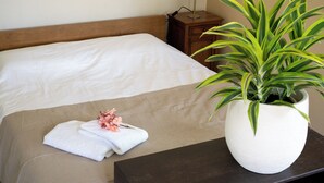 Un letto rifatto con cura, con una coperta beige, un cuscino bianco e un piccolo bouquet di fiori sul comodino. Accanto al letto, una pianta in vaso su un altro comodino.