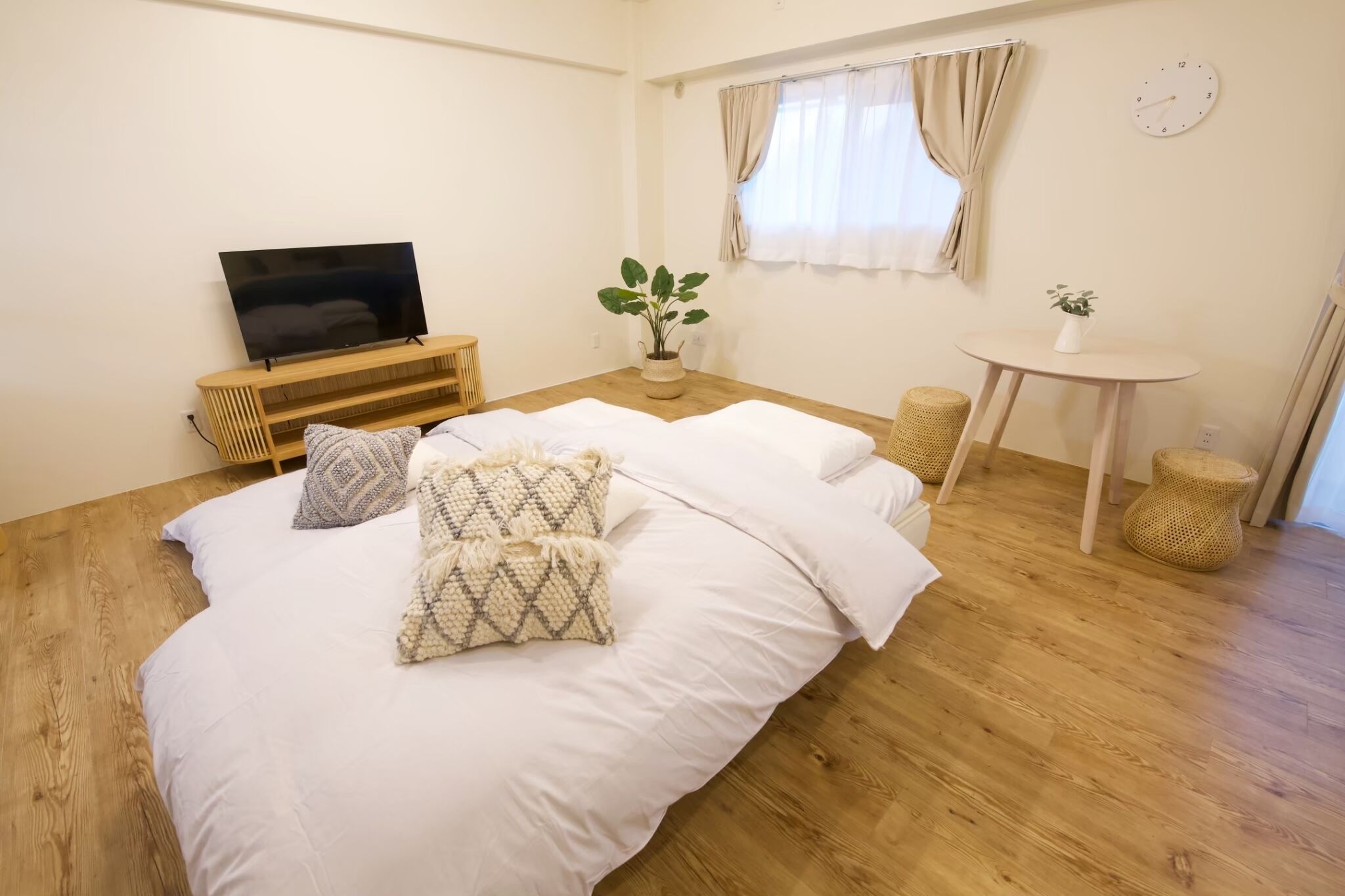 A bedroom with a bed, a TV on a wooden stand, a small table with a plant, and a window with curtains.