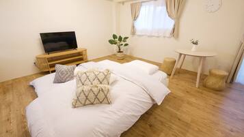 A bedroom with a bed, a TV on a wooden stand, a small table with a plant, and a window with curtains.