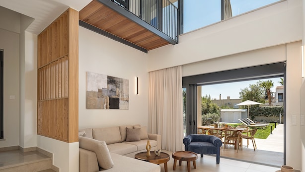 Una sala de estar moderna con un sofá seccional, una pequeña mesa redonda y un sillón azul. Hay una escalera que sube a un nivel superior, y un amplio ventanal corredero se abre a un patio exterior con un comedor y sillas de descanso.