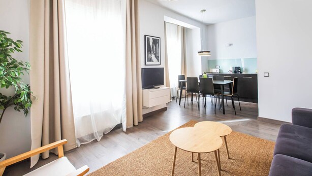 A modern living room with a sofa, a coffee table, and a TV. There is a dining area with chairs and a table, and a kitchen with a counter and appliances.