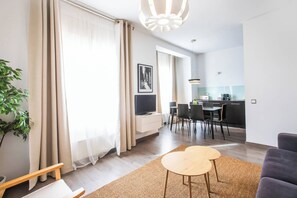 A modern living room with a sofa, a coffee table, and a TV. There is a dining area with chairs and a table, and a kitchen with a counter and appliances.