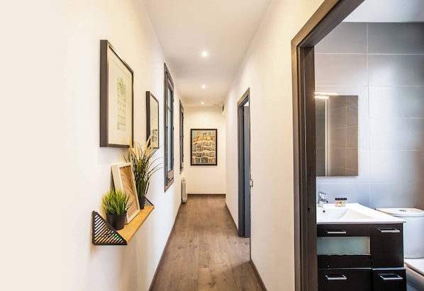 A hallway with framed artwork on the walls, a wooden shelf with a plant, and a modern bathroom with a large mirror and white sink.
