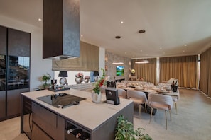 Une cuisine moderne dotée d’un îlot central, d’un coin repas avec une table dressée pour six personnes et d’une grande fenêtre agrémentée de rideaux.