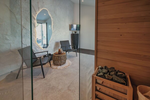 A modern interior with two black chairs, a round wooden table, a large mirror, a television on the wall, and a wooden bench with rocks. - Villa Kim Momjan With Pool and Spa (Buje)