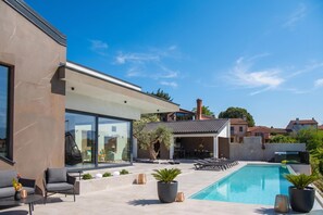 Een modern buitenzwembad met een glazen patio, lounge fauteuils en potplanten.