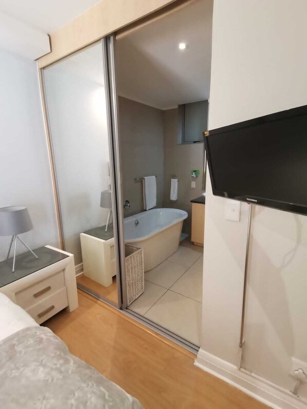 Un baño moderno con bañera, inodoro y lavamanos. Hay un televisor montado en la pared y un espejo sobre el lavamanos.