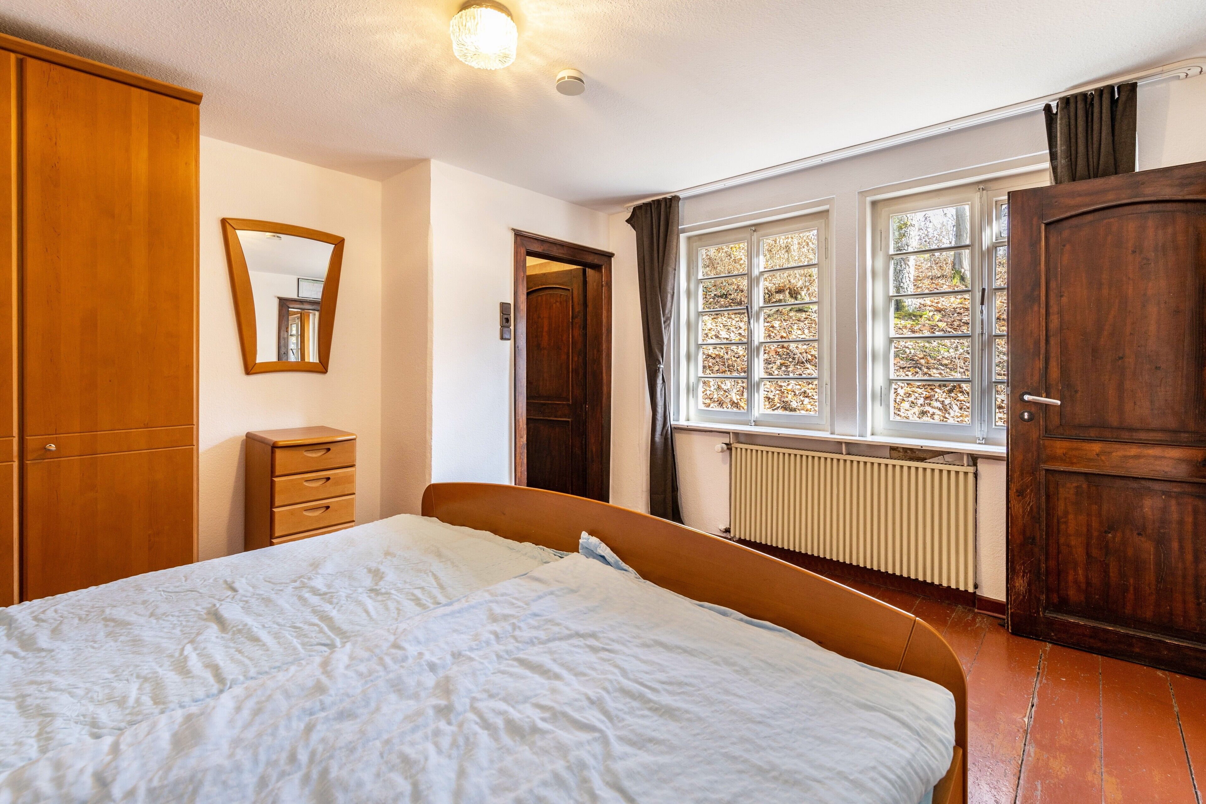 Een slaapkamer met een bed, een houten garderobe, een dressoir, een spiegel en een raam met gordijnen.