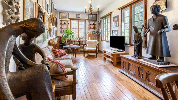 A room with wooden flooring, a sofa, a chair, a television, and various sculptures and paintings on the walls. - Holiday Home 'Hueseken' with Shared Garden and Wi-Fi (Finnentrop)