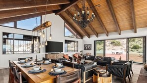 Un vaste espace de salle à manger avec une table en bois dressée pour le repas, un canapé en cuir noir et une grande fenêtre donnant sur l’extérieur.