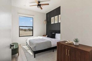 Une chambre moderne avec une grande fenêtre, un lit recouvert d’une housse de couette à carreaux gris et blancs, une tête de lit en bois, une table de chevet en bois, un pouf vert et une commode en bois sur laquelle est posée une plante.