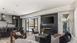 A modern living room with a sofa, coffee table, and chairs. There is a kitchen area with a dining table and chairs. A large flat-screen TV is mounted on the wall.