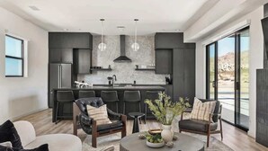 A modern living room with a large window, a kitchen with dark cabinetry, and a seating area with leather chairs.