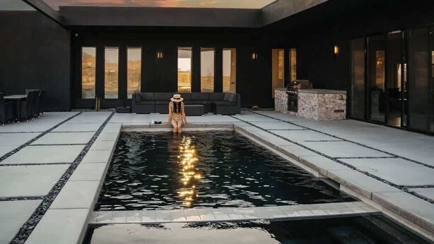 A modern outdoor pool area with a person sitting by the pool, surrounded by large windows and a minimalist design.