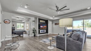 Ein geräumiges Wohnzimmer mit grauem Sofa, einer Poolbillardkugel, einem Deckenventilator und einem großen Fenster mit Blick ins Freie.