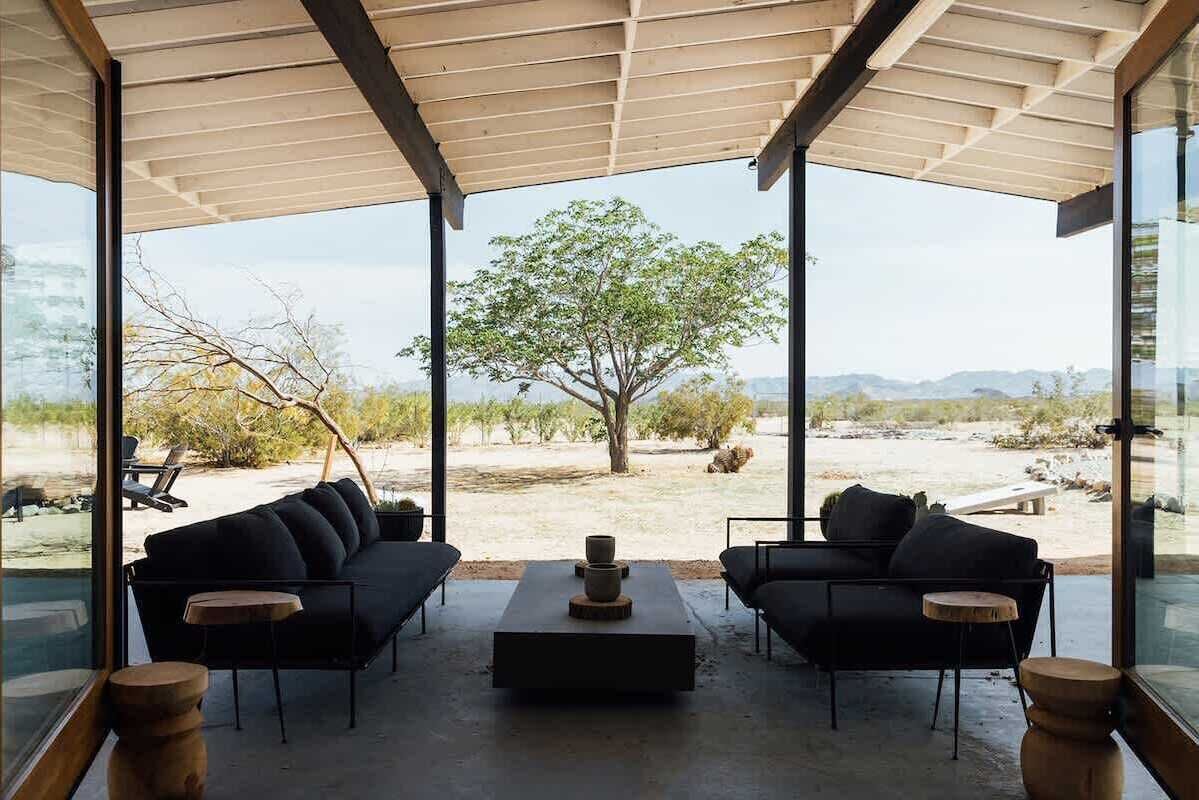 A modern outdoor seating area with a sofa and coffee table, large windows, and a view of a tree and landscape.