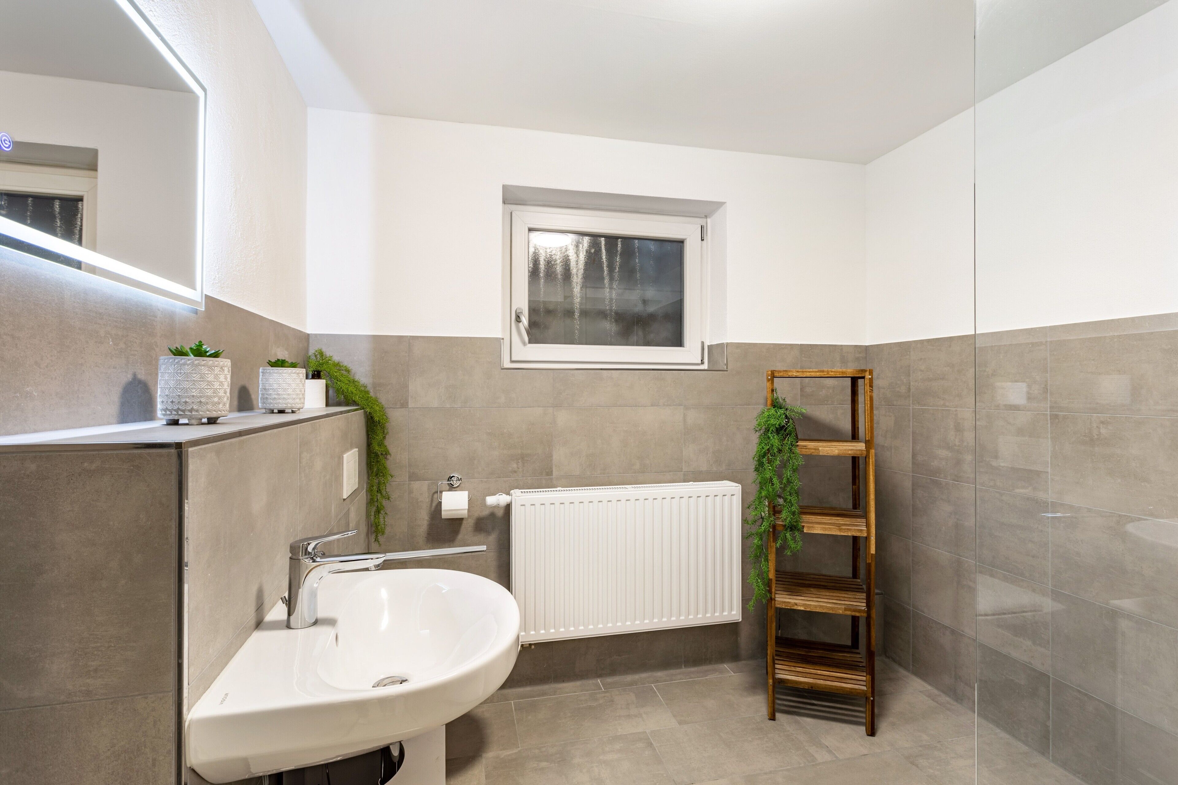 Ein Badezimmer mit einem weißen Waschbecken, einem Fenster, einer Heizung und einem Holzbodenregal mit Pflanzen.