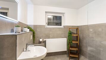 Ein Badezimmer mit einem weißen Waschbecken, einem Fenster, einer Heizung und einem Holzbodenregal mit Pflanzen.