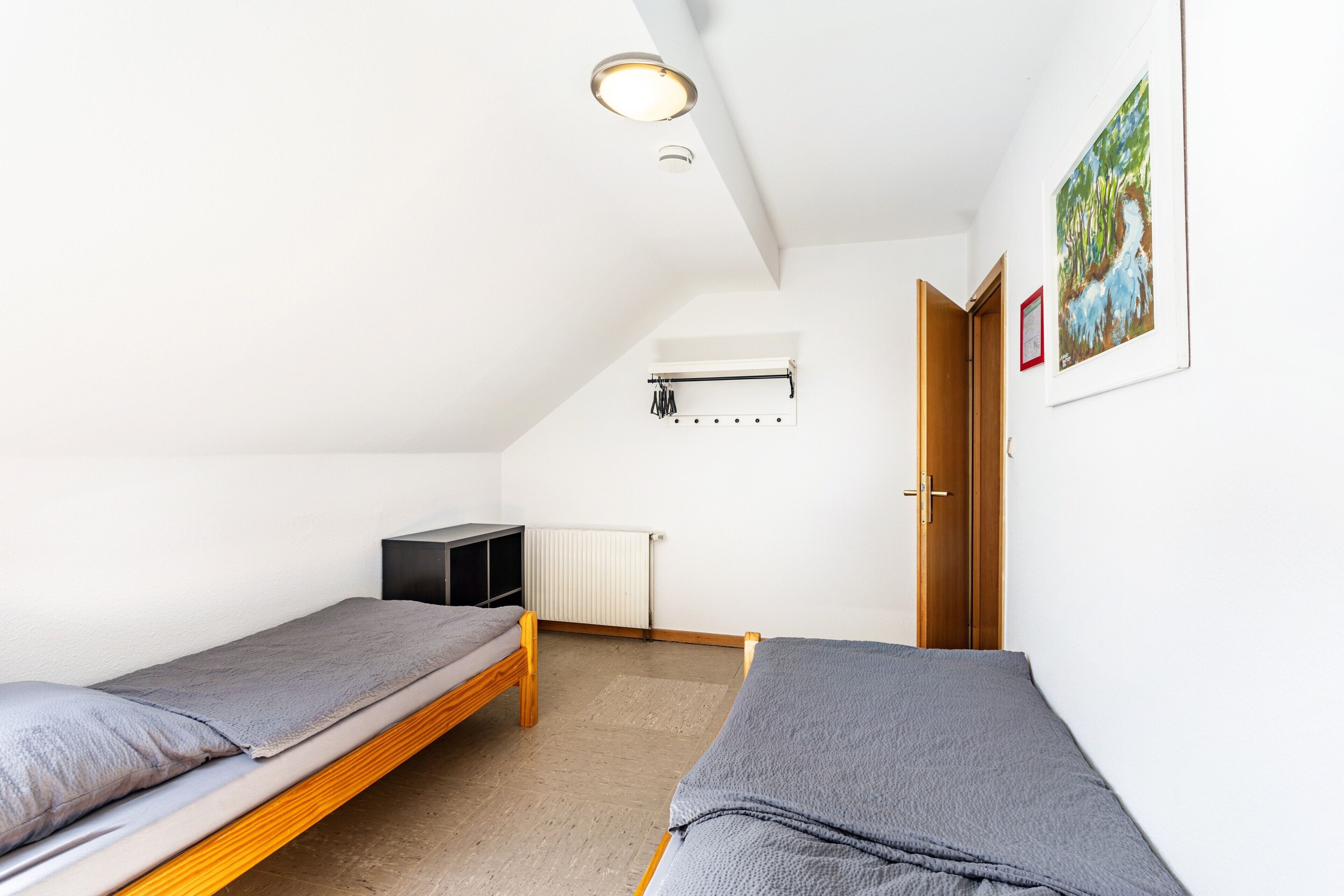 Een kleine, lichte kamer met twee eenpersoonsbedden, een radiator, een houten deur en een schilderij in een lijst aan de muur.