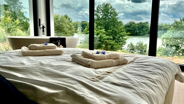 A bedroom with a large bed, two towels, a bathtub, and a view of a lake and trees. - Amazing villa with swimming pool next to the lake. (Wrzeszewo)