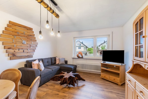 A modern living room with a sectional sofa, a wooden wall clock, a flat-screen TV, and a wooden dining table.