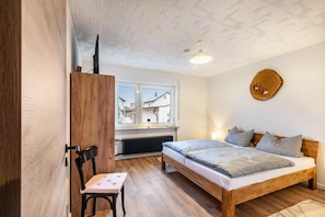 A bedroom with a wooden bed, a chair, a radiator, and a window with a view of houses.