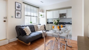A modern living room with a grey sofa, a small dining table with a clear chair, and a kitchen area with white cabinets and a microwave. - Classic 1BR - Notting Hill Gem (London)