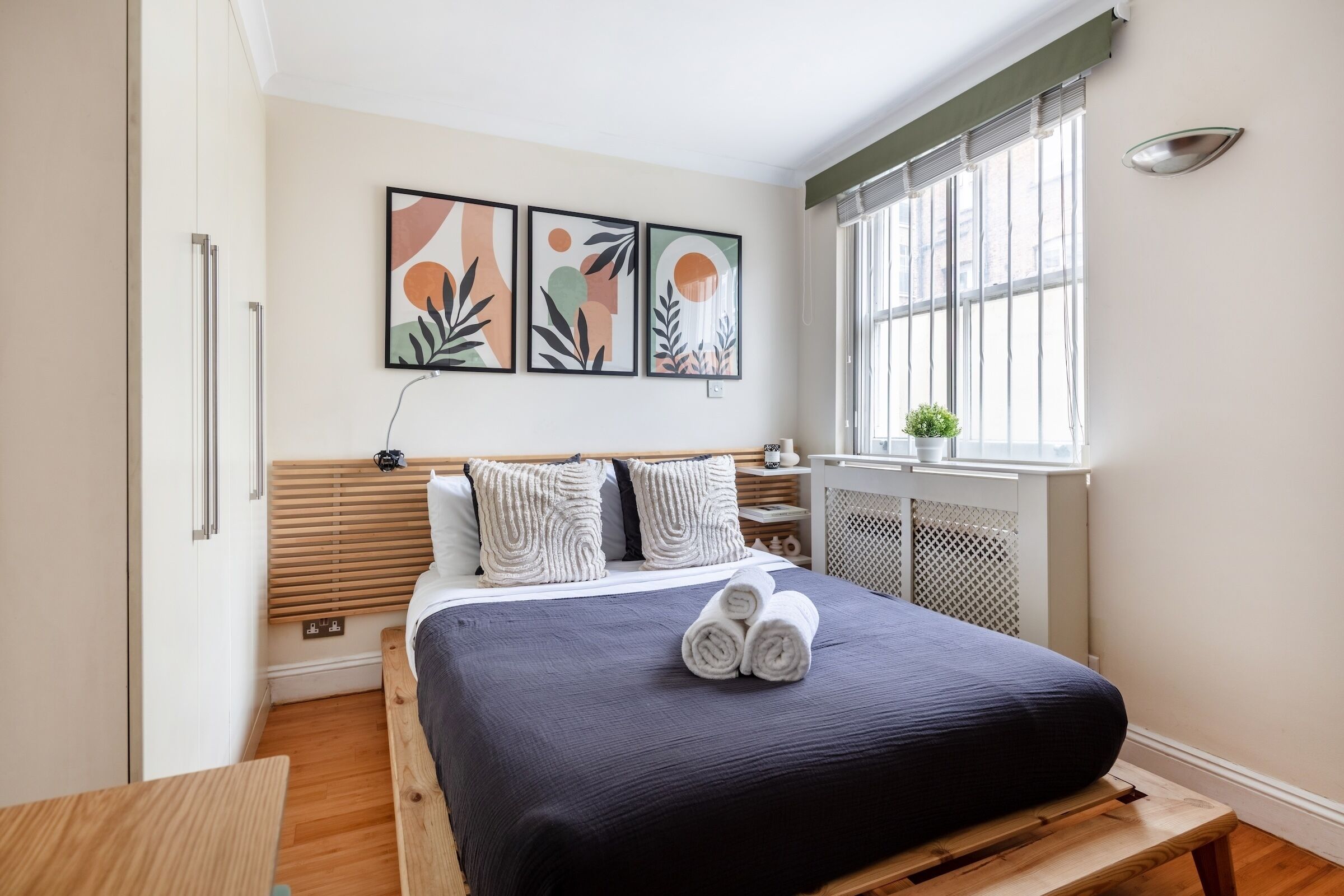 Een moderne slaapkamer met een groot bed, een nachtkastje, een raam met jaloezieën en abstracte wanddecoratie.
