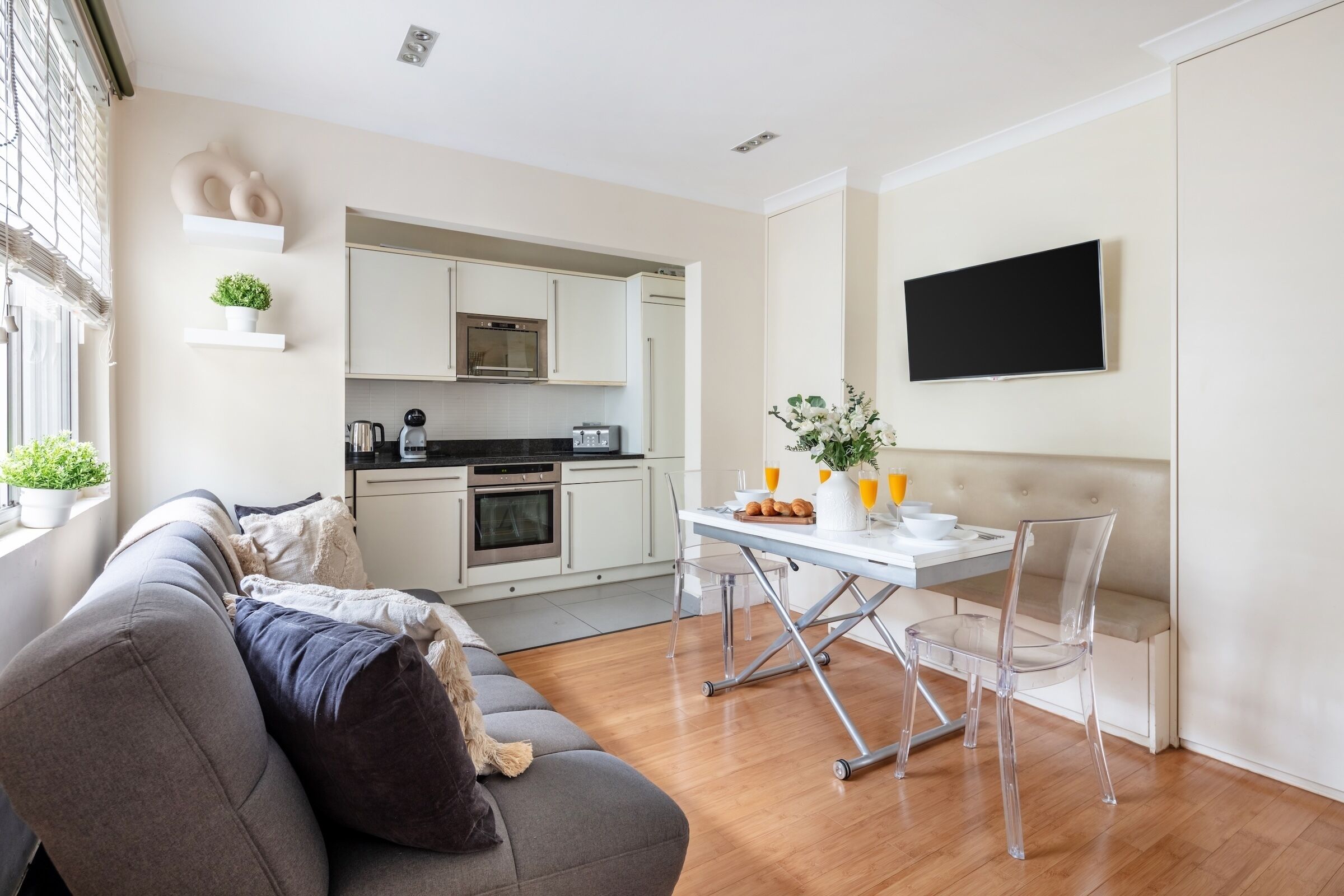Un salon moderne avec un canapé gris, une table à manger avec des chaises, une cuisine en arrière-plan et un téléviseur fixé au mur.
