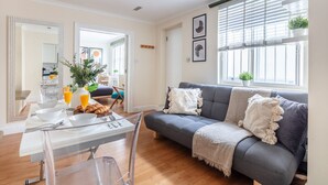 A modern living room with a grey sofa, a dining table with breakfast items, and a large window with blinds. - Classic 1BR - Notting Hill Gem (London)
