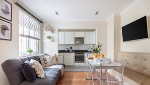 A modern living space with a grey sofa, a dining table with clear chairs, a kitchen area, and a flat-screen TV mounted on the wall. - Classic 1BR - Notting Hill Gem (London)