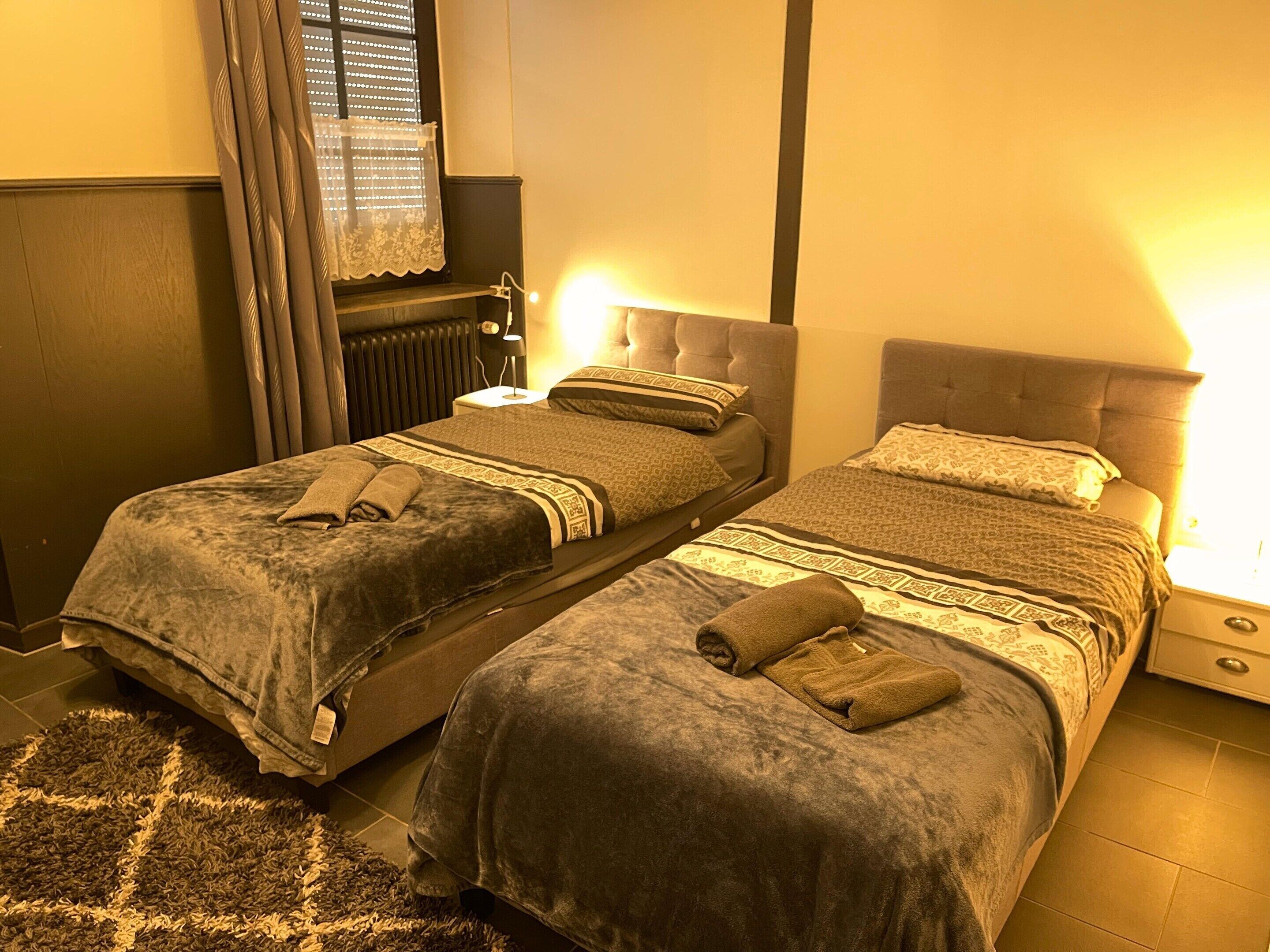 A twin room with two beds, a radiator, a window with curtains, and a nightstand with a lamp.