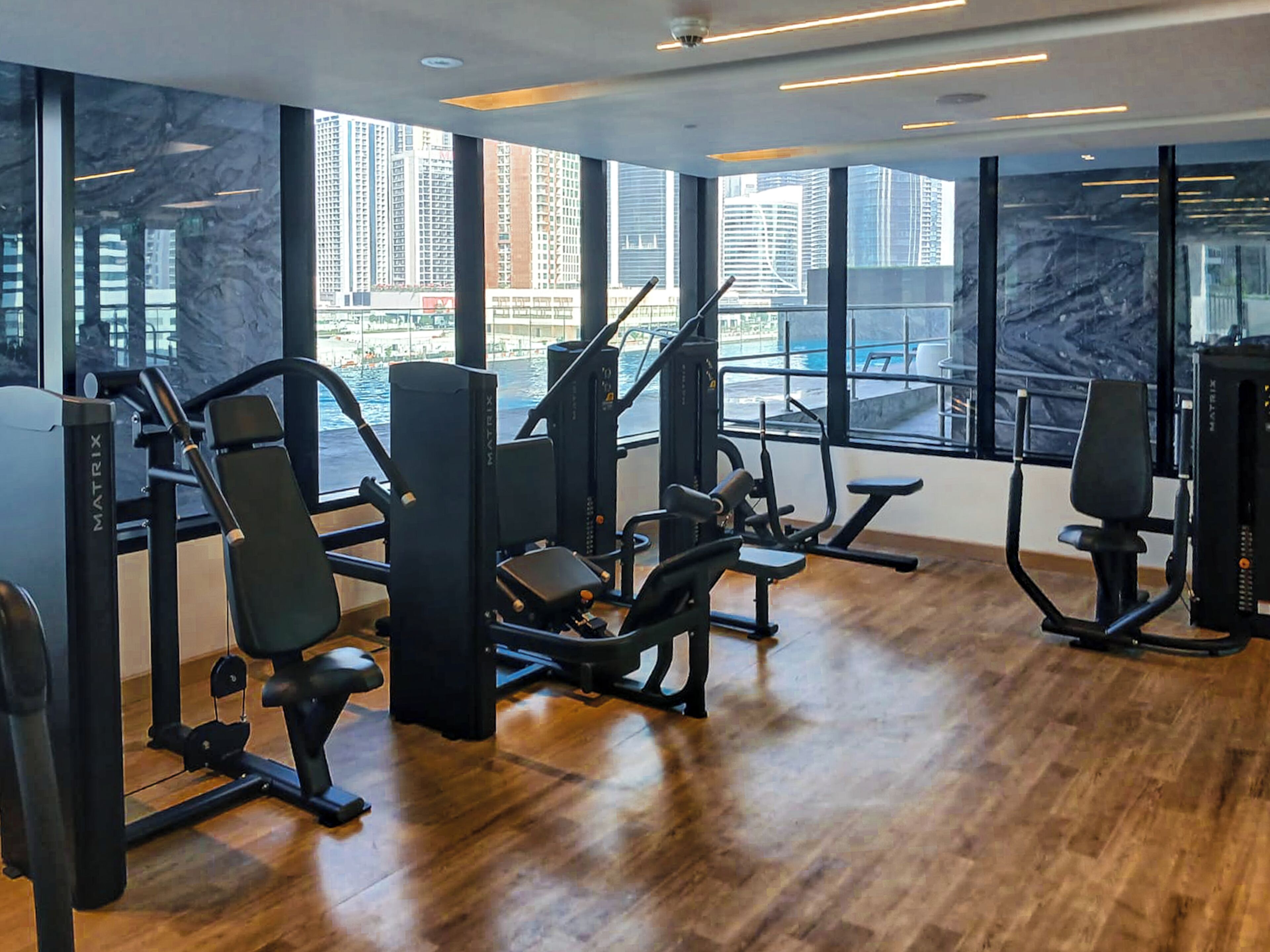 A modern gym with various exercise machines, large windows offering city views, and a polished wooden floor.