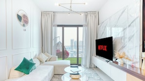 A modern living room with a sofa, a coffee table, a flat-screen TV, and a view of the cityscape through large windows. - PVH - Exquisite 1BR - Binghatti Canal (Dubai)